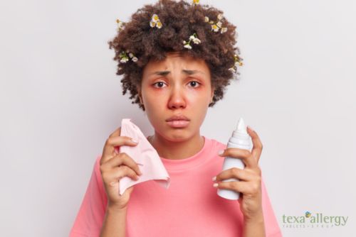 Disappointed young Afro American woman uses nasal aerosol suffers from allergic rhinitis has red swollen eyes looks unhappily at camera dressed in casual t shirt, with Texa Allergy Logo in the bottom right corner.